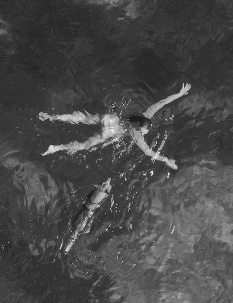 A woman and her kind dog swim together in the crystal waters of the Dalmatian seaside. The sea surface is glistening in the intense summer sunshine, the water is turquoise and clear.