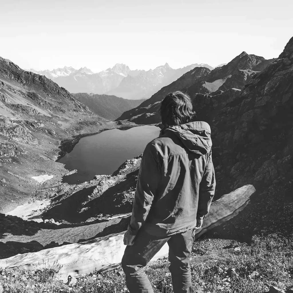 Explorer standing on a mountainside, overlooking a serene mediterranean lake surrounded by lush, green valleys and rugged mountain peaks.